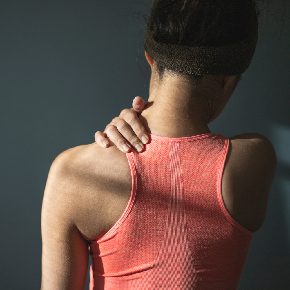 Person seen from behind wearing a coral athletic tank top while rubbing or massaging their neck/shoulder with one hand on the opposite shoulder, indoors.