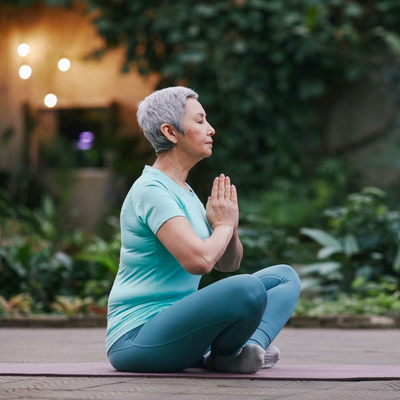 Older woman with short gray hair sits cross-legged on a pink yoga mat in a garden, hands in prayer.