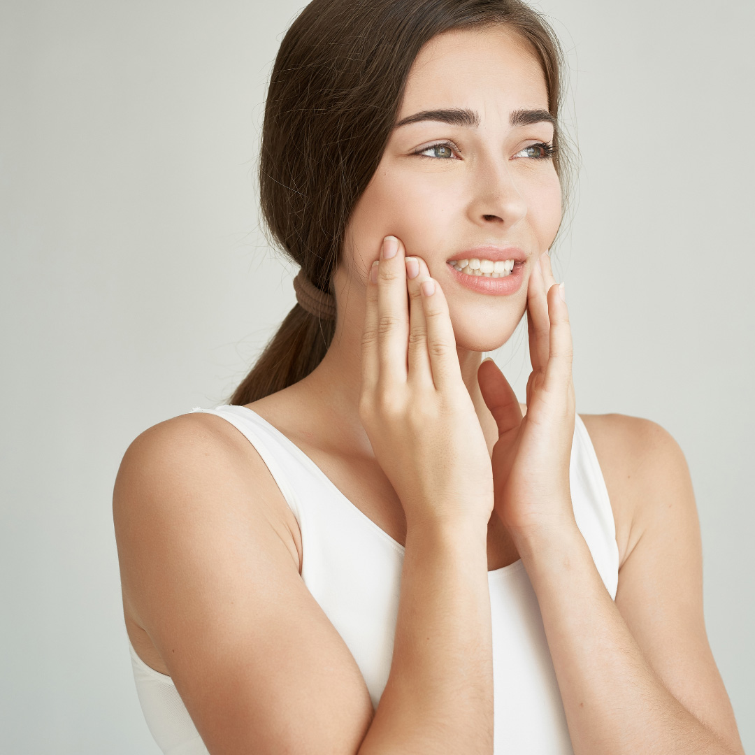Woman in a white tank top gently pressing her cheeks with both hands, smiling thoughtfully at the camera's left side.
