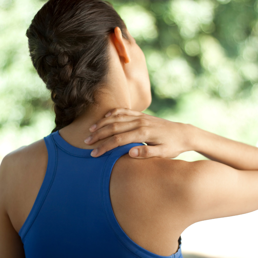 Person in a blue sleeveless top holds the back of their neck, suggesting neck strain or stretching outdoors.
