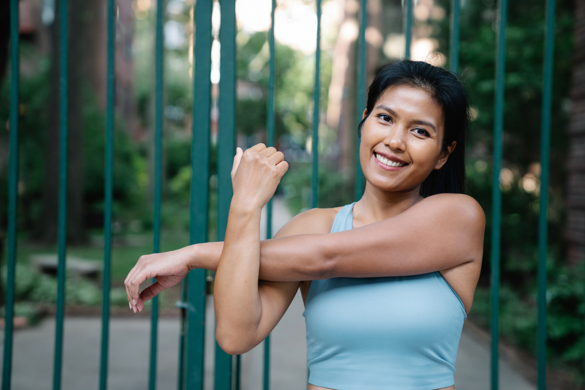 Smiling woman in a light blue athletic top stretches her arms outdoors beside a green metal fence.