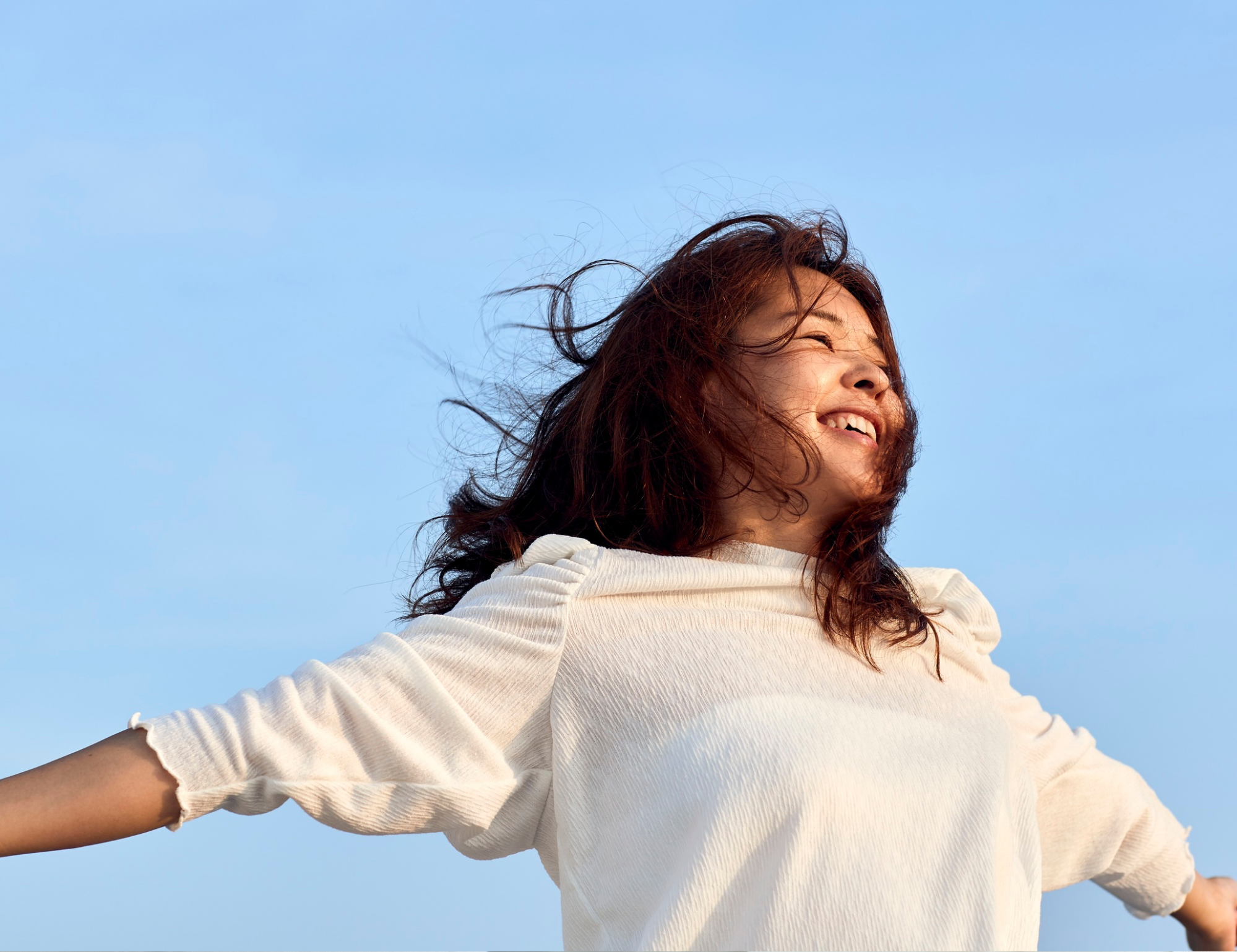Woman smiling with wind-swept hair against a clear blue sky, wearing a white sweater.