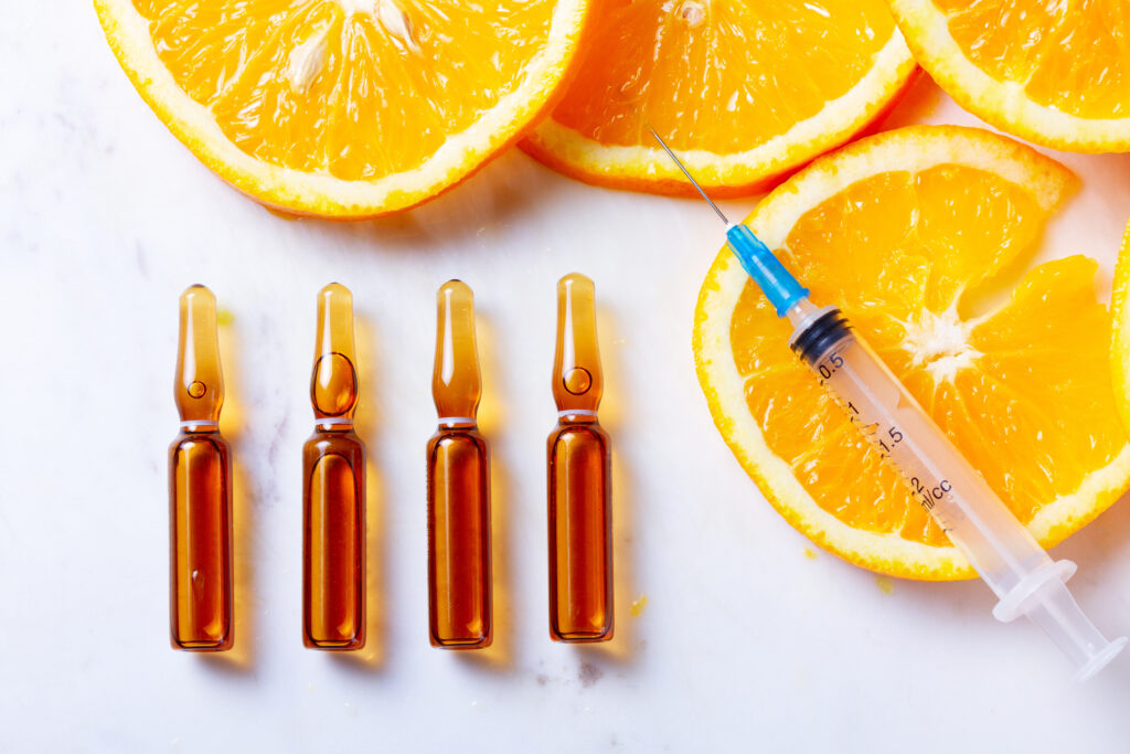 Syringe and four amber glass vials lined up on a white surface with orange halves in the background, suggesting a medical or vitamin-shot scene.