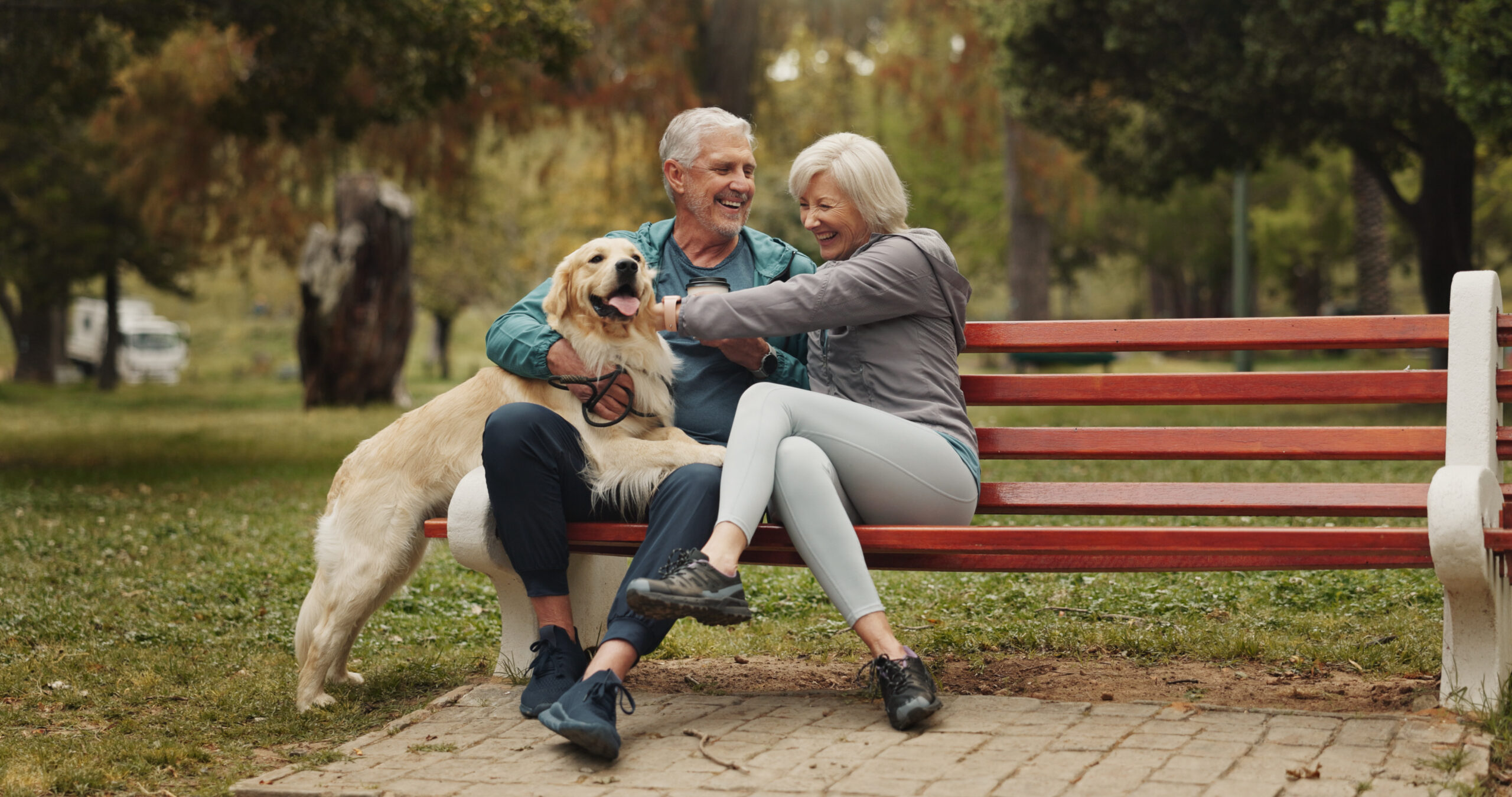 Senior couple sitting on a red park bench, smiling as their golden retriever stands beside them on a leash in a leafy park.