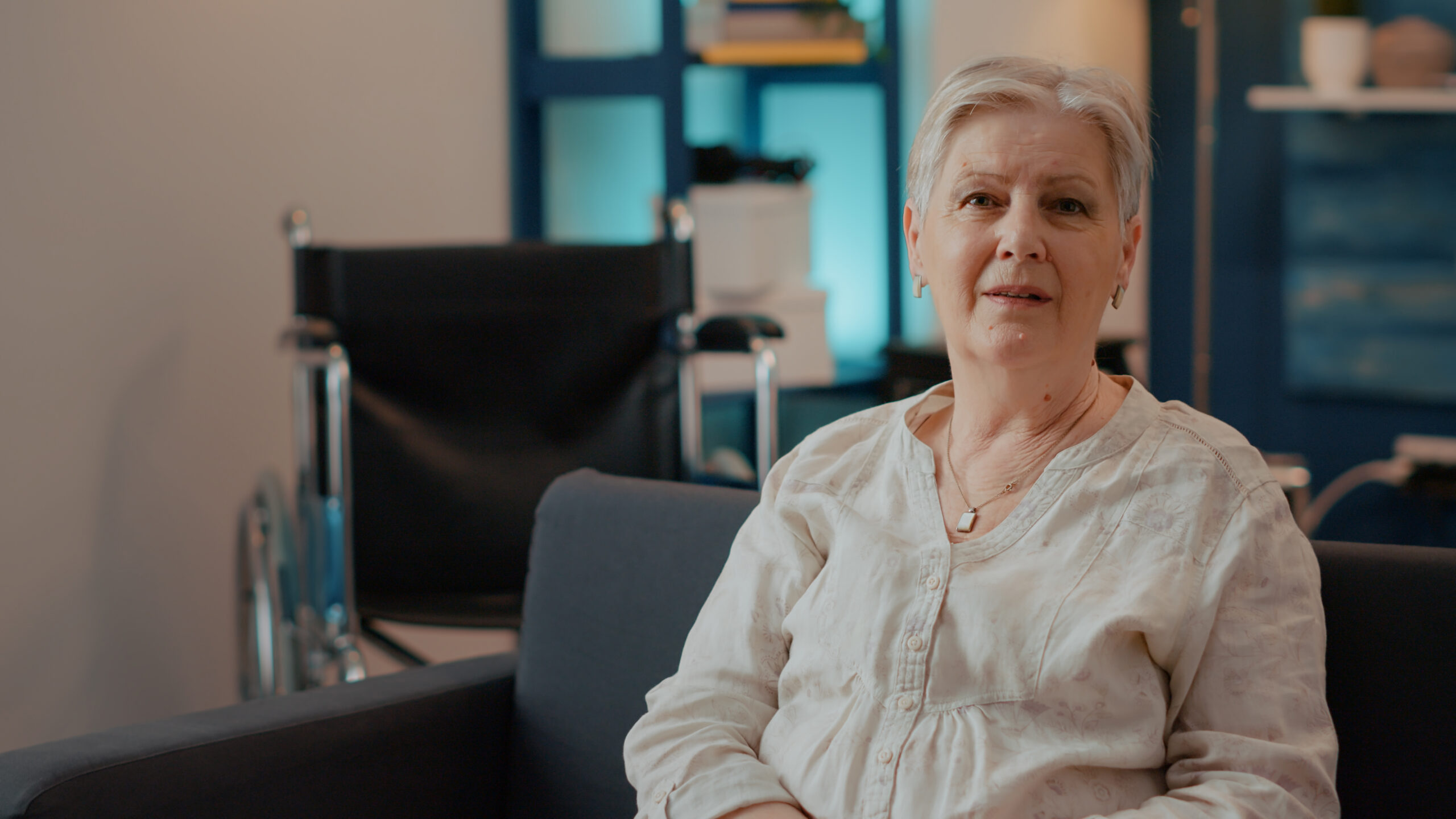 Elderly woman with short gray hair in a light blouse seated in a clinic waiting area, with a wheelchair in the background.